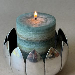 May include: A cylindrical pillar candle with horizontal bands of blue, green, and white wax, featuring a lit wick with a small orange flame. The candle is held within a polished silver-toned metal candle holder shaped like a blooming lotus flower.
