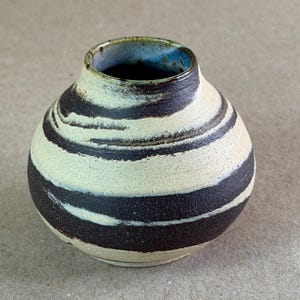 May include: A small, round ceramic vase featuring a textured surface with horizontal, swirling bands of cream and dark brown. The interior of the vase is glazed in a deep blue, and the rim shows a subtle, dark brown edge finish.