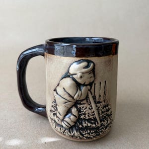 May include: A ceramic mug with a dark brown handle and rim. The mug features a relief sculpture of a person leaning on a fence. The mug is beige with brown accents, and the handle is a dark brown color.