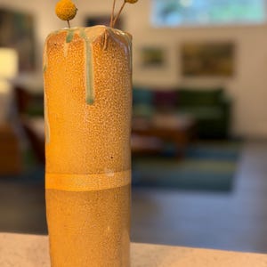 May include: A tall, cylindrical ceramic vase in shades of yellow and brown, with a textured surface. The vase has a band of lighter color around the middle and green glaze drips at the top. It holds several dried, yellow, spherical flowers.