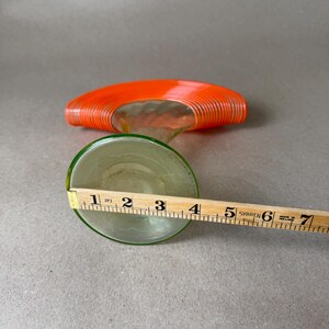 May include: A clear glass decorative object with an orange, spiral-shaped top and a green-rimmed base. The base is approximately 4 inches in diameter. The object is on a gray surface.