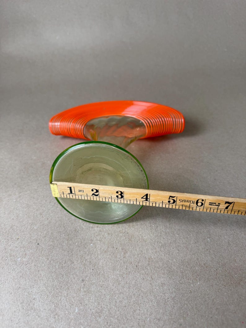 May include: A clear glass decorative object with an orange, spiral-shaped top and a green-rimmed base. The base is approximately 4 inches in diameter. The object is on a gray surface.