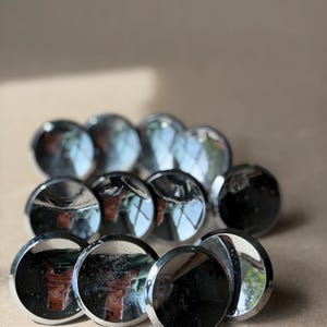 May include: A collection of shiny, circular, chrome-colored cabinet knobs. The knobs are arranged in a cluster, reflecting light and the surrounding environment. The knobs have a smooth, polished surface.