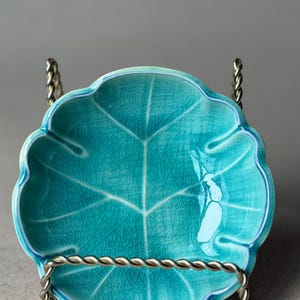 May include: A turquoise ceramic leaf-shaped dish with a detailed vein pattern. The dish is displayed on a silver-colored metal stand with a twisted rope design. The dish has a scalloped edge and a glossy finish.