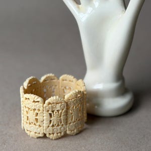 May include: A cream-colored cuff bracelet composed of multiple rectangular links, each featuring intricate, symmetrical openwork carvings of floral motifs and geometric patterns. The bracelet has a continuous, repeating design.