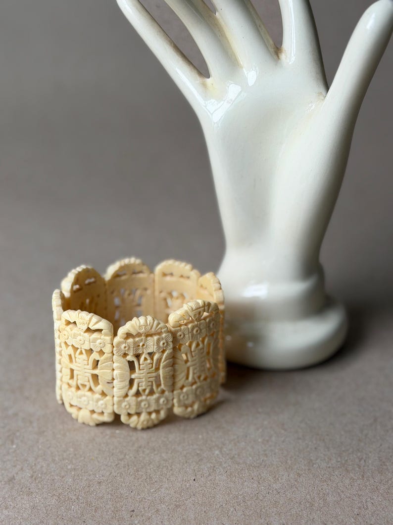 May include: A cream-colored cuff bracelet composed of multiple rectangular links, each featuring intricate, symmetrical openwork carvings of floral motifs and geometric patterns. The bracelet has a continuous, repeating design.
