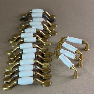 May include: A collection of drawer pulls featuring a white ceramic center handle with polished brass metal ends. The set includes multiple curved handles arranged in a long row and a smaller group of four matching pulls positioned to the side.