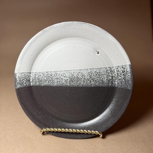 May include: A handmade ceramic plate with a two-tone design. The top half is white, the bottom half is dark gray, and a speckled gray band separates the two colors. The plate is displayed on a gold-colored metal stand.