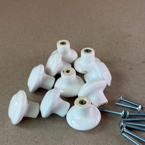 May include: A collection of nine white ceramic cabinet knobs with a smooth, rounded mushroom-shaped design. Each knob features a central metal threaded insert on the underside for mounting. Several silver-colored metal screws are included separately.