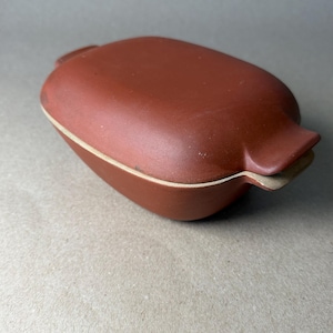 May include: A reddish-brown ceramic container with a lid. The container has an oval shape with rounded edges and a light-colored rim. The lid fits snugly, and the container appears to be handmade.