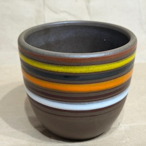 May include: A cylindrical ceramic cup with a matte brown base and a series of horizontal stripes in yellow, black, orange, and white encircling the upper exterior. The interior of the cup is a dark brown, and the rim is smooth and rounded.