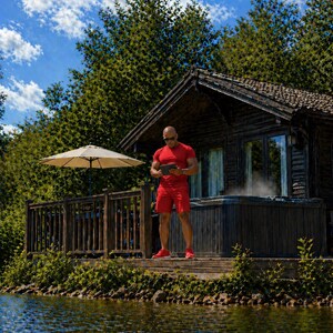 May include: A man in red athletic wear stands on a wooden deck near a lake, holding a tablet. A wooden cabin with a hot tub and a beige umbrella are visible. The sky is blue with fluffy white clouds, and the water reflects the surroundings.