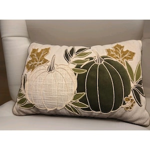 May include: A rectangular throw pillow with a cream-colored herringbone fabric base. The design features an embroidered white textured pumpkin and a dark green velvet pumpkin, accented with embroidered gold and green leaves and stems.