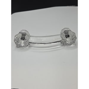 May include: A clear glass cabinet pull with two faceted, flower-shaped knobs connected by a curved, clear glass bar. The hardware is designed for a cabinet or drawer.