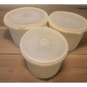 May include: Three off-white plastic food storage containers with matching lids. The containers are cylindrical with rounded edges. The lids have a circular design in the center. The containers are arranged on a wooden surface.