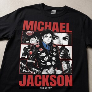 May include: Black t-shirt with a graphic of Michael Jackson. The design features the name "MICHAEL JACKSON" in red, a portrait of Jackson, and the text "KING OF POP" below the image.
