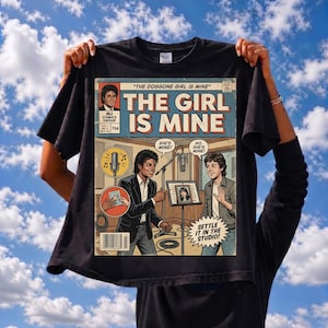Pode incluir: Camiseta preta com uma ilustração em estilo de história em quadrinhos com o texto "THE GIRL IS MINE". A arte retrata dois homens em um estúdio de gravação, com balões de fala e o texto "SETTLE IT IN THE STUDIO!". A camiseta é exibida contra um céu azul.