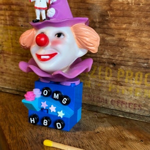 May include: A purple and white clown head made of plastic with a red nose and a big smile. The clown head is on top of a blue Lego block with the letters OMG HBD on it. There are three pink and white stars on the block. A small clown figurine is standing on top of the clown head.