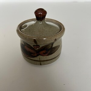 May include: A small, ceramic jar with a lid. The jar is a speckled tan color with a brown floral design. The lid has a small, rounded handle. The jar is likely used for storing spices or other small items.