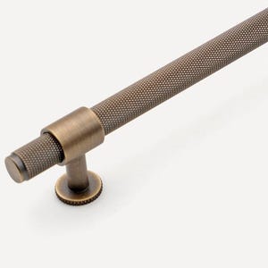 May include: A bronze-toned cabinet pull with a textured, cylindrical handle and matching round end caps. The pull is mounted on two small, round posts. The logo "GOLDEN HOME HARDWARE" is in the upper left corner.
