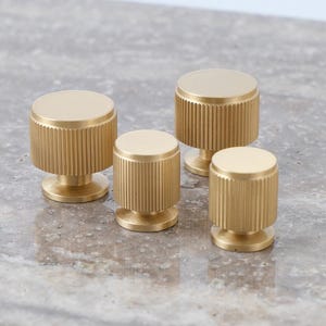 以下が含まれることがあります： 溝付き円筒形デザインの4つのゴールドカラーのキャビネットノブ。ノブは円形のベースと平らな上面を持っています。「GOLDEN HOME HARDWARE」のロゴが左上にあります。