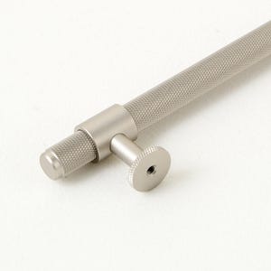 Puede incluir: Un tirador de armario plateado con un mango cilíndrico texturizado y un pequeño pomo redondo. El herraje presenta una textura moleteada en el mango y los extremos del pomo. El logotipo "GOLDEN HOME HARDWARE" está en la esquina superior izquierda.