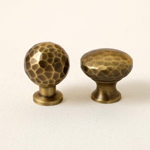 May include: Two antique brass cabinet knobs. One is a round, hammered-texture knob, and the other is an oval knob with a similar hammered texture. The knobs are set against a white background. The logo "GOLDEN HOME HARDWARE" is in the upper left corner.