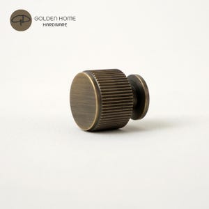 May include: A bronze-colored cabinet knob with a ridged cylindrical body and a circular base. The knob has a dark finish and is set against a white background. The logo "GOLDEN HOME HARDWARE" is in the upper left corner.