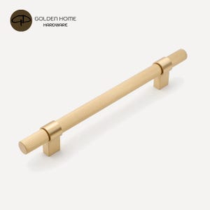 K&ouml;nnte beinhalten: Ein goldfarbener Schrankgriff mit geriffeltem, zylindrischem Griff und quadratischen Montagepfosten. Die Hardware tr&auml;gt den Text "GOLDEN HOME HARDWARE" und ein Logo. Der Griff ist f&uuml;r M&ouml;bel und Schr&auml;nke konzipiert.