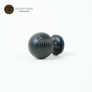 K&ouml;nnte beinhalten: Ein schwarzer Schrankknauf mit einem runden, geriffelten Griff und einem glatten, kreisf&ouml;rmigen Sockel. Der Knauf ist aus Metall gefertigt und f&uuml;r den Einsatz an M&ouml;beln konzipiert. Das Logo "GOLDEN HOME HARDWARE" befindet sich in der oberen linken Ecke.