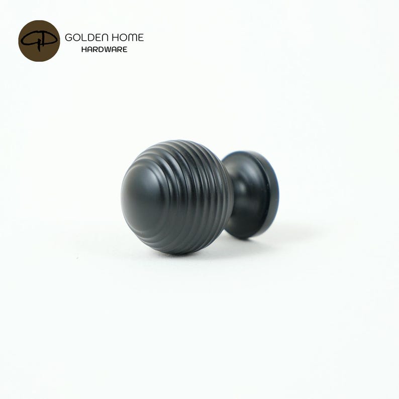 K&ouml;nnte beinhalten: Ein schwarzer Schrankknauf mit einem runden, geriffelten Griff und einem glatten, kreisf&ouml;rmigen Sockel. Der Knauf ist aus Metall gefertigt und f&uuml;r den Einsatz an M&ouml;beln konzipiert. Das Logo "GOLDEN HOME HARDWARE" befindet sich in der oberen linken Ecke.