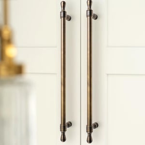 Op de afbeelding: Twee bronskleurige kastgrepen met een verticaal, cilindrisch ontwerp. Elke greep heeft een decoratieve boven- en onderkant en is gemonteerd op een witte kastdeur. Het logo "GOLDEN HOME HARDWARE" staat in de linkerbovenhoek.