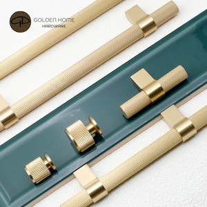 Puede incluir: Varias piezas de herrajes dorados para armarios, incluyendo tiradores y pomos, están dispuestas sobre una superficie turquesa. Los herrajes tienen una textura acanalada y un diseño moderno. El logotipo "GOLDEN HOME HARDWARE" es visible.