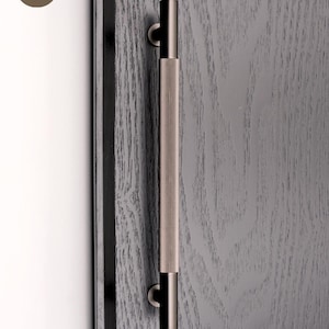 Może przedstawiać: Zbliżenie drzwi z ciemnoszarym wykończeniem z usłojeniem drewna. Pionowo wyśrodkowana klamka do drzwi ze szczotkowanego metalu. Klamka ma żebrowaną część środkową i cylindryczne końce. Logo Golden Home Hardware znajduje się w lewym górnym rogu.