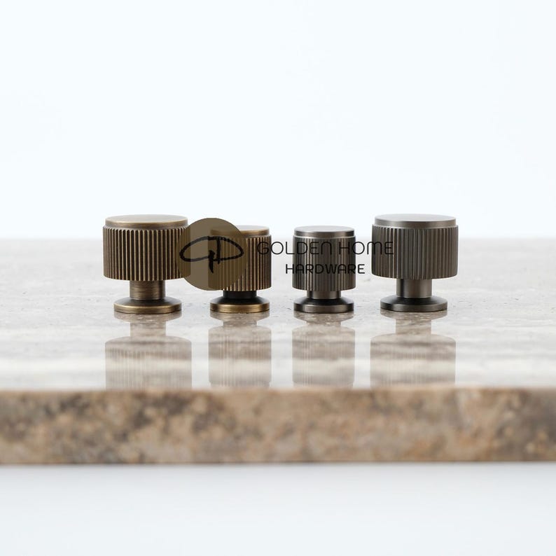 May include: Four metal cabinet knobs with a ribbed design. The knobs are in shades of bronze and gray, with a circular top and a short, cylindrical base. The knobs are arranged in a row on a light brown surface. The text "GOLDEN HOME HARDWARE" is visible.