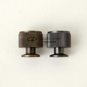 May include: Two metal cabinet knobs with a ridged design. One knob is bronze-colored with a logo, and the other is dark gray. The knobs have a cylindrical shape with a short, round base. The text "GOLDEN HOME HARDWARE" is visible.