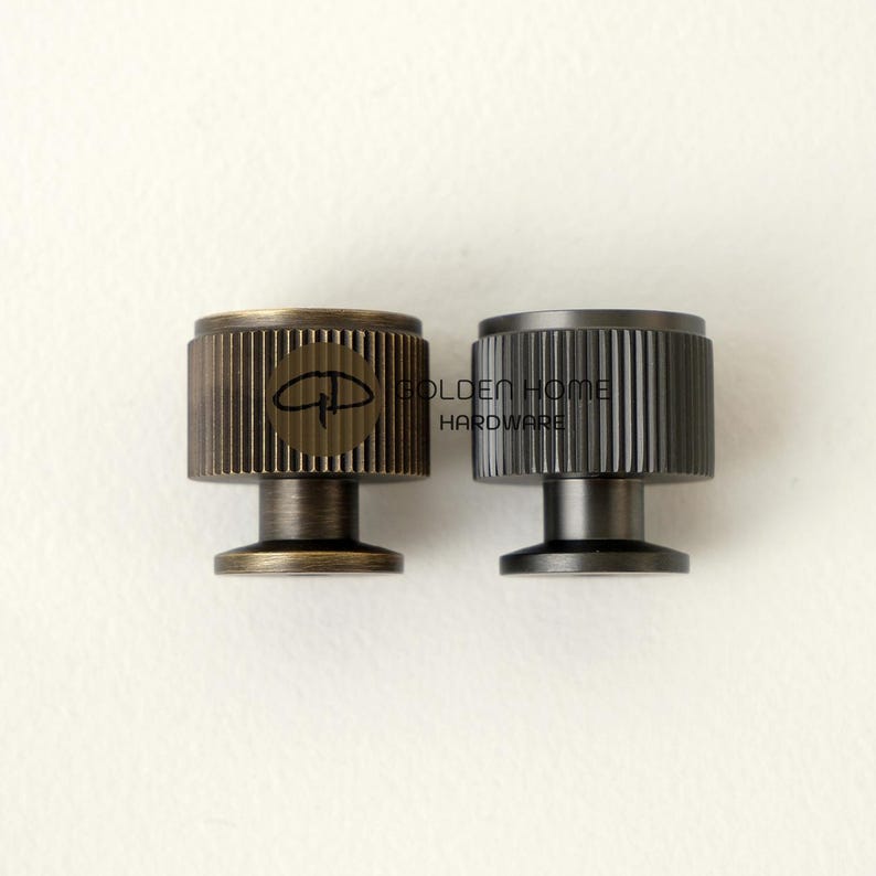 May include: Two metal cabinet knobs with a ridged design. One knob is bronze-colored with a logo, and the other is dark gray. The knobs have a cylindrical shape with a short, round base. The text "GOLDEN HOME HARDWARE" is visible.
