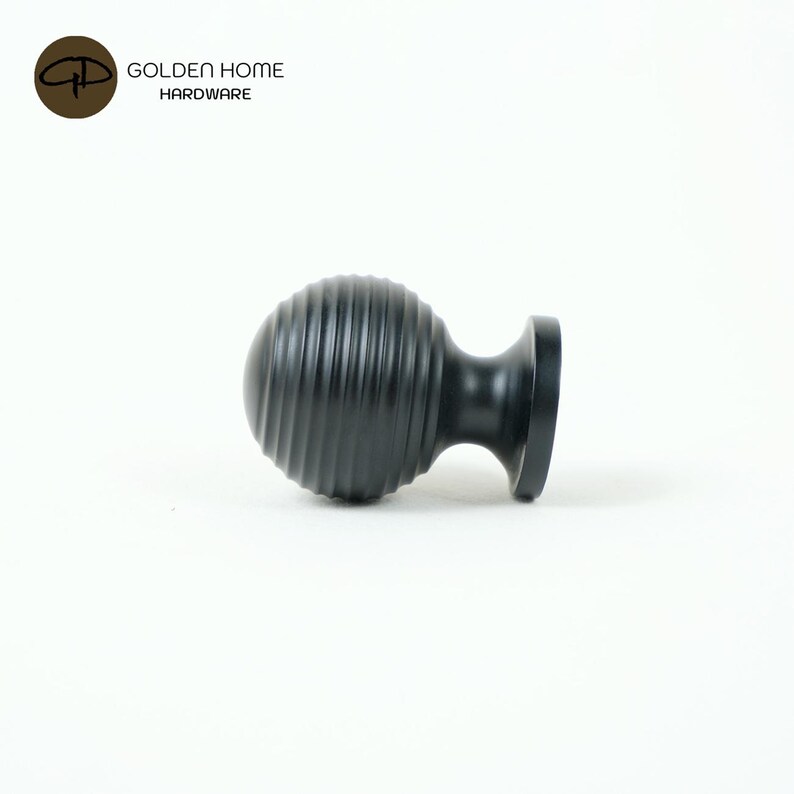 K&ouml;nnte beinhalten: Ein schwarzer, geriffelter Schrankknauf mit einer runden, kugelartigen Form und einem flachen Sockel. Der Knauf ist aus Metall und hat eine matte Oberfl&auml;che. Das Logo "GOLDEN HOME HARDWARE" befindet sich in der oberen linken Ecke.