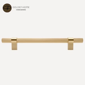 K&ouml;nnte beinhalten: Ein goldfarbener Schrankgriff mit geriffeltem, zylindrischem Griff und quadratischen Befestigungspfosten. Die Hardware hat eine geb&uuml;rstete Oberfl&auml;che und ist f&uuml;r eine einfache Installation konzipiert. Das Logo "GOLDEN HOME HARDWARE" befindet sich in der oberen linken Ecke.