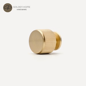 K&ouml;nnte beinhalten: Ein goldfarbener Schrankknopf mit geriffeltem zylindrischen K&ouml;rper und flacher, kreisf&ouml;rmiger Fl&auml;che. Der Knopf ist aus Metall und hat eine glatte, polierte Oberfl&auml;che. Das Logo "GOLDEN HOME HARDWARE" befindet sich oben links.