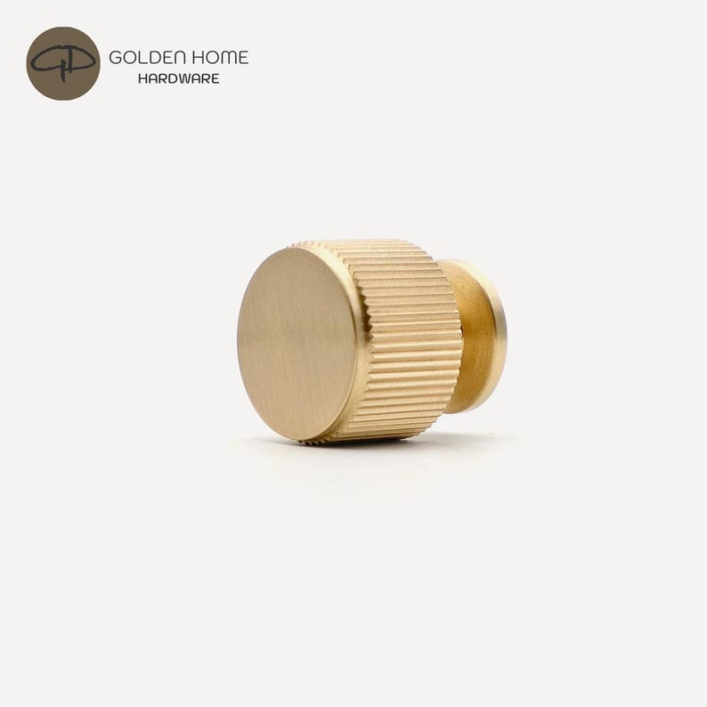 K&ouml;nnte beinhalten: Ein goldfarbener Schrankknopf mit geriffeltem zylindrischen K&ouml;rper und flacher, kreisf&ouml;rmiger Fl&auml;che. Der Knopf ist aus Metall und hat eine glatte, polierte Oberfl&auml;che. Das Logo "GOLDEN HOME HARDWARE" befindet sich oben links.