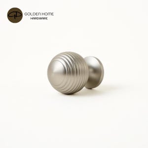 K&ouml;nnte beinhalten: Zwei Schrankkn&ouml;pfe aus geb&uuml;rstetem Nickel. Ein Knopf hat ein geriffeltes, kugelf&ouml;rmiges Design, der andere ist eine glatte Kugel. Das Logo "GOLDEN HOME HARDWARE" befindet sich in der oberen linken Ecke.