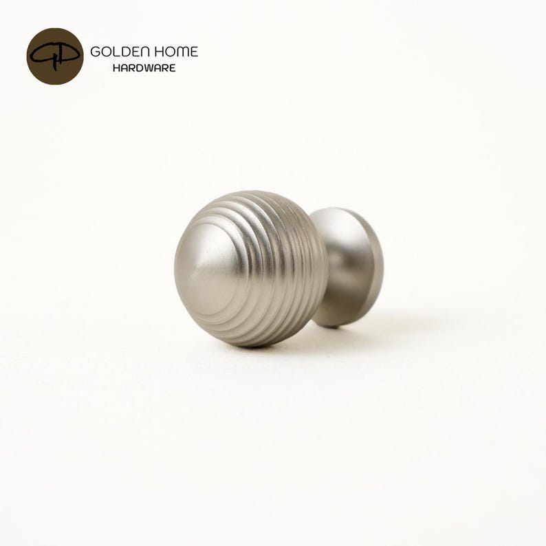 K&ouml;nnte beinhalten: Zwei Schrankkn&ouml;pfe aus geb&uuml;rstetem Nickel. Ein Knopf hat ein geriffeltes, kugelf&ouml;rmiges Design, der andere ist eine glatte Kugel. Das Logo "GOLDEN HOME HARDWARE" befindet sich in der oberen linken Ecke.