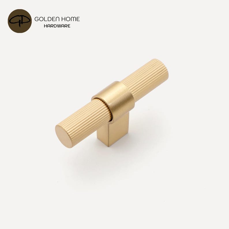 K&ouml;nnte beinhalten: Ein goldfarbener Schrankgriff mit einem zylindrischen, geriffelten Griff und einem quadratischen Sockel. Der Griff ist mit einem glatten, kreisf&ouml;rmigen Band mit dem Sockel verbunden. Der Text "GOLDEN HOME HARDWARE" befindet sich in der oberen linken Ecke.