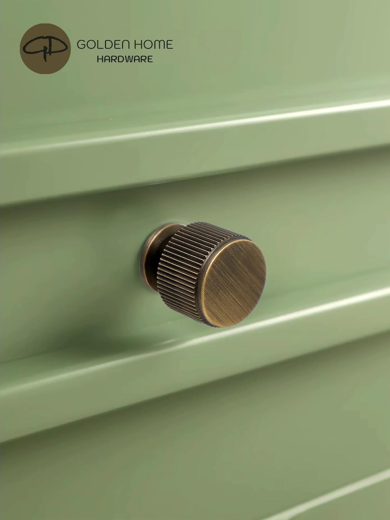May include: Close-up of a bronze cabinet knob with a ridged design, mounted on a light green cabinet drawer. The knob has a circular shape and a vintage aesthetic. The text "GOLDEN HOME HARDWARE" is visible in the upper left corner.
