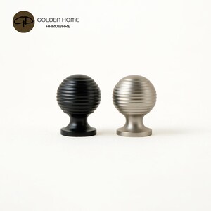 K&ouml;nnte beinhalten: Zwei dekorative Schrankkn&ouml;pfe. Einer ist schwarz, der andere silberfarben. Beide haben ein rundes, geripptes Design und einen kurzen Sockel. Die Kn&ouml;pfe sind von Golden Home Hardware.