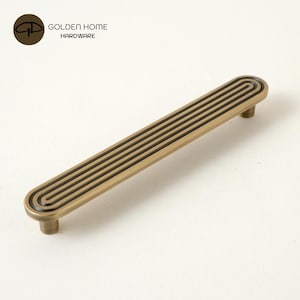 May include: A brass-toned cabinet pull with a grooved design. The rectangular handle features a series of parallel lines and rounded ends. The hardware is from Golden Home Hardware.