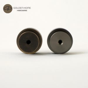 May include: Two metal cabinet knobs. One is a dark bronze color, and the other is a silver color. Both knobs have a circular design with a ridged edge and a central hole. The knobs are from Golden Home Hardware.