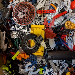 Op de afbeelding: Een wirwar van kleurrijke plastic speelgoedonderdelen, waaronder tandwielen, wielen en diverse structurele componenten. De stukken zijn in tinten rood, geel, zwart, zilver en groen. Een cirkelvormig stuk met een doolhofontwerp is zichtbaar.