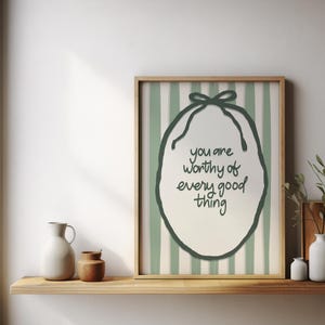 May include: A framed print with the text "you are worthy of every good thing" in a whimsical font. The artwork features a green oval shape with a bow at the top, set against a striped background. Decorative items sit on a wooden shelf.